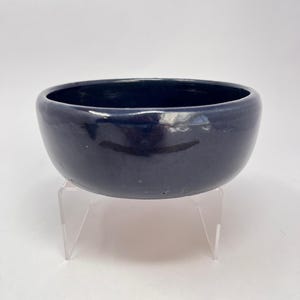 Peut inclure: Un bol en céramique bleu marine avec une finition brillante. Le bol est rond avec une large ouverture et repose sur un support en acrylique transparent. L'intérieur du bol est d'une nuance de bleu plus foncée.