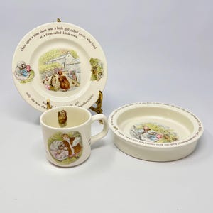 Puede incluir: Un juego de vajilla infantil de color crema con ilustraciones y texto de un libro de cuentos. El juego incluye un plato, una taza y un cuenco pequeño. El plato y el cuenco son circulares, la taza tiene un asa.