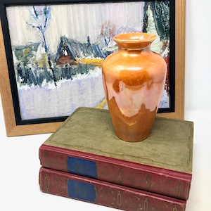 Op de afbeelding: Een oranje iriserende vaas staat op twee antieke boeken met rode en groene covers. Op de achtergrond een ingelijst landschapsschilderij met een winterse scène, met een besneeuwd landschap en een houten lijst.