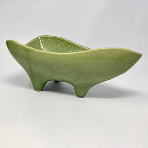 May include: A mid-century modern, green ceramic bowl with a unique, elongated shape. The bowl has three small, rounded feet and gently curved sides. The interior has a textured surface, and the exterior has a smooth, matte finish. A decorative piece.