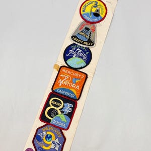 Puede incluir: Un conjunto vintage de parches de la misión Mercury de la NASA sobre un respaldo de cartón. Los parches presentan diseños coloridos con textos como "Freedom 7", "Liberty Bell 7" y "Friendship 6". La etiqueta de precio dice $3.50.