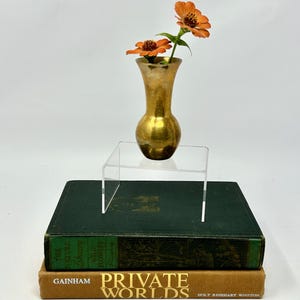 May include: A small, gold-colored vase holding two orange flowers, displayed on a clear acrylic stand. The stand rests on a stack of two vintage books with green and tan covers. The books are titled "The Nature Library" and "Private Worlds."