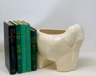 Puede incluir: Un macetero de cerámica blanca con forma de elefante con una pila de libros verdes detrás. Los libros tienen títulos como "Foxfire", "Under the Eye of the Storm" y "India Allan".