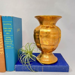 Puede incluir: Un jarrón dorado con una superficie texturizada, colocado sobre un libro azul. El jarrón tiene una base ancha y una parte superior acampanada. Una pequeña planta aérea y dos libros están al lado del jarrón.