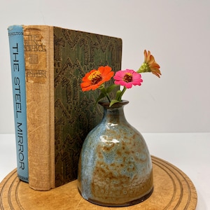 Puede incluir: Un pequeño jarrón de cerámica azul y marrón contiene dos flores coloridas. El jarrón está sobre una bandeja de madera junto a dos libros antiguos. Un libro tiene una columna vertebral azul con el título "THE STEEL MIRROR".