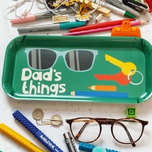 K&ouml;nnte beinhalten: Ein gr&uuml;nes Tablett mit den Worten "Dad's things" darauf gedruckt. Das Tablett hat eine Sonnenbrille, einen Schl&uuml;ssel und einen Bleistift darauf gezeichnet. Auf einer wei&szlig;en Oberfl&auml;che um das Tablett herum befinden sich verschiedene Gegenst&auml;nde, darunter Brillen, Stifte, Bleistifte und eine Batterie.
