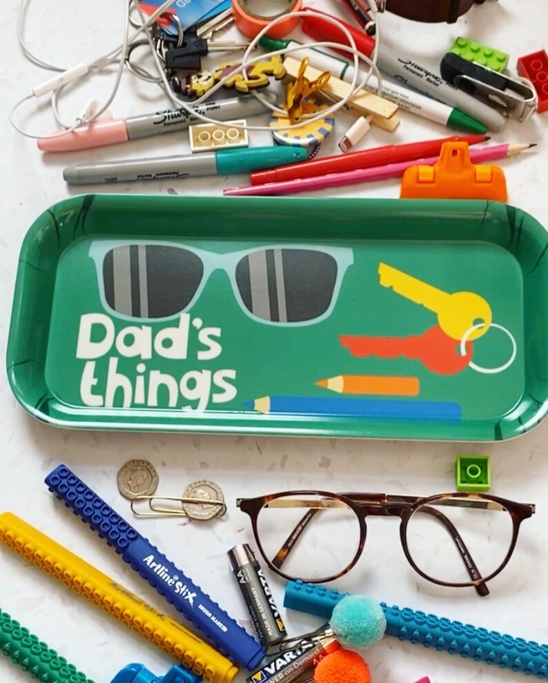 K&ouml;nnte beinhalten: Ein gr&uuml;nes Tablett mit den Worten "Dad's things" darauf gedruckt. Das Tablett hat eine Sonnenbrille, einen Schl&uuml;ssel und einen Bleistift darauf gezeichnet. Auf einer wei&szlig;en Oberfl&auml;che um das Tablett herum befinden sich verschiedene Gegenst&auml;nde, darunter Brillen, Stifte, Bleistifte und eine Batterie.
