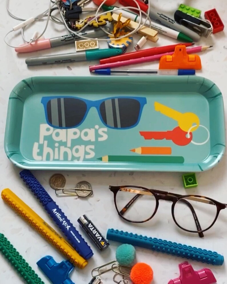 K&ouml;nnte beinhalten: Ein blaues Tablett mit dem Text "Papa's things" und einer Sonnenbrille, einem Schl&uuml;ssel und einem Bleistift darauf. Das Tablett ist von verschiedenen Gegenst&auml;nden umgeben, darunter eine Brille, ein gelber Stift, ein blauer Stift, eine Batterie und ein paar andere Gegenst&auml;nde.