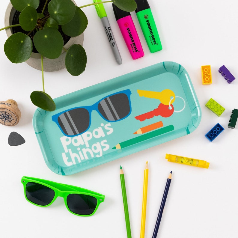 K&ouml;nnte beinhalten: Ein blaues Tablett mit dem Text "Papa's things" und einer Sonnenbrille, Schl&uuml;sseln und Bleistiften. Das Tablett ist von bunten Bleistiften, Bausteinen und Markern umgeben.