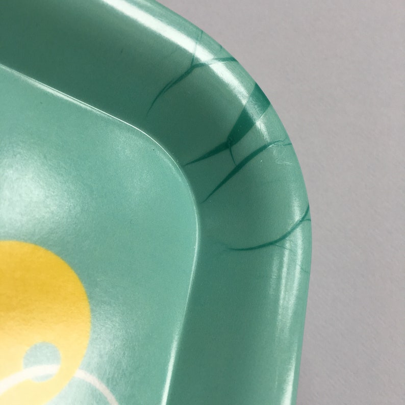 K&ouml;nnte beinhalten: Ein t&uuml;rkisfarbenes Tablett aus Metall mit einem gelben und wei&szlig;en Design. Das Tablett hat ein paar Kratzer auf der Oberfl&auml;che.