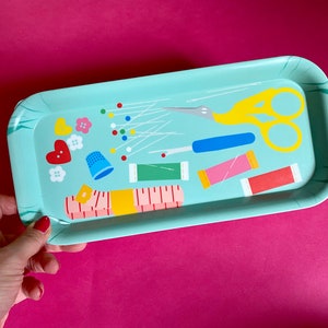 May include: A rectangular, pale blue tray decorated with sewing-related illustrations. The tray shows yellow scissors, spools of thread, a thimble, pins, and a measuring tape. The tray is held by a person with red nail polish.