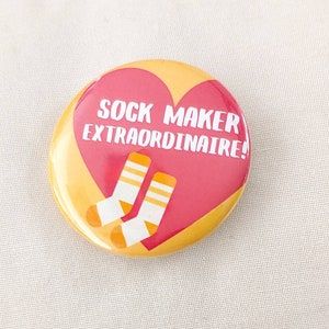 Puede incluir: Un botón amarillo y rojo con forma de corazón con el texto "SOCK MAKER EXTRAORDINAIRE!" y dos calcetines blancos con rayas amarillas.