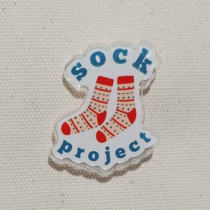 May include: A white acrylic pin with a blue outline and the words "sock project" in blue letters. Two red and white striped socks with polka dots are pictured above the words.