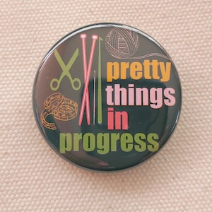 May include: A black button with a green background and the text "pretty things in progress". The button features a variety of craft supplies including scissors, yarn, needles, and a tape measure.