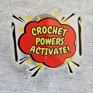 May include: A red and white comic book style speech bubble with the text "CROCHET POWERS ACTIVATE!" inside. The speech bubble is surrounded by yellow stars and black and white lightning bolts. Two black crochet hooks are on either side of the speech bubble.