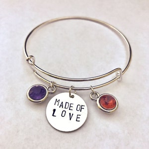 May include: A silver bangle bracelet with three charms: a purple gemstone, a red gemstone, and a silver charm with the words "MADE OF LOVE" engraved on it.