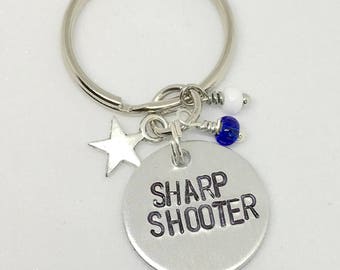 May include: Silver keychain with a star charm, blue and white beads, and a round tag that says "SHARP SHOOTER".