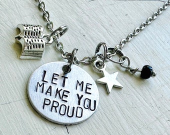 May include: A silver-toned necklace with multiple charms. The charms include a stack of books, a star, a black bead, and a round pendant with the phrase "LET ME MAKE YOU PROUD" engraved on it. The chain is delicate and the overall design is simple.