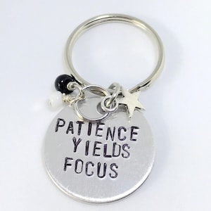 May include: Silver keychain with a round charm that says "Patience Yields Focus". The charm is attached to a silver key ring with a small silver star and black and white beads.