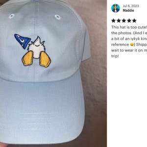 May include: A light blue baseball cap with an embroidered image of a cartoon duck wearing a blue wizard hat. The duck is facing away from the viewer and has its wings outstretched.