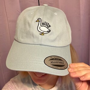 May include: A light blue baseball cap with an embroidered white duck and the word "silly". The cap has an adjustable strap with a brown label that says "The Authentic Yupoong Adjustable Yupoong Since 1974".