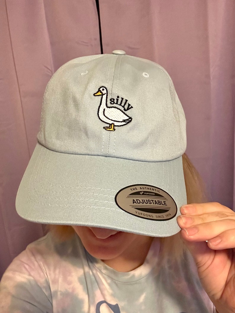 May include: A light blue baseball cap with an embroidered white duck and the word "silly". The cap has an adjustable strap with a brown label that says "The Authentic Yupoong Adjustable Yupoong Since 1974".