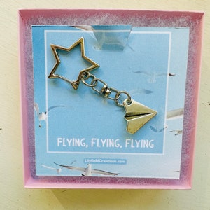 May include: A silver keychain featuring a star charm and a paper aeroplane charm. The keychain is presented in a pink box with a blue background and the text "FLYING, FLYING, FLYING" and "LilyfieldCreations.com".