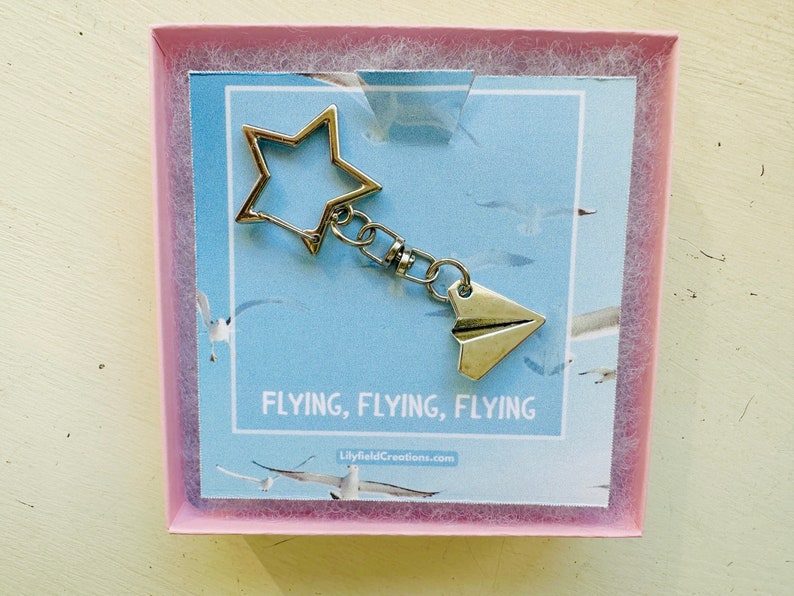 May include: A silver keychain featuring a star charm and a paper aeroplane charm. The keychain is presented in a pink box with a blue background and the text "FLYING, FLYING, FLYING" and "LilyfieldCreations.com".