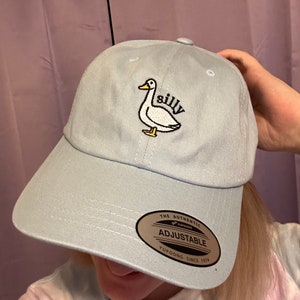May include: A light blue baseball cap with an embroidered white duck and the word "silly" on it. The cap has an adjustable strap with a brown label that says "The Authentic Yupoong Adjustable Yupoong Since 1974."
