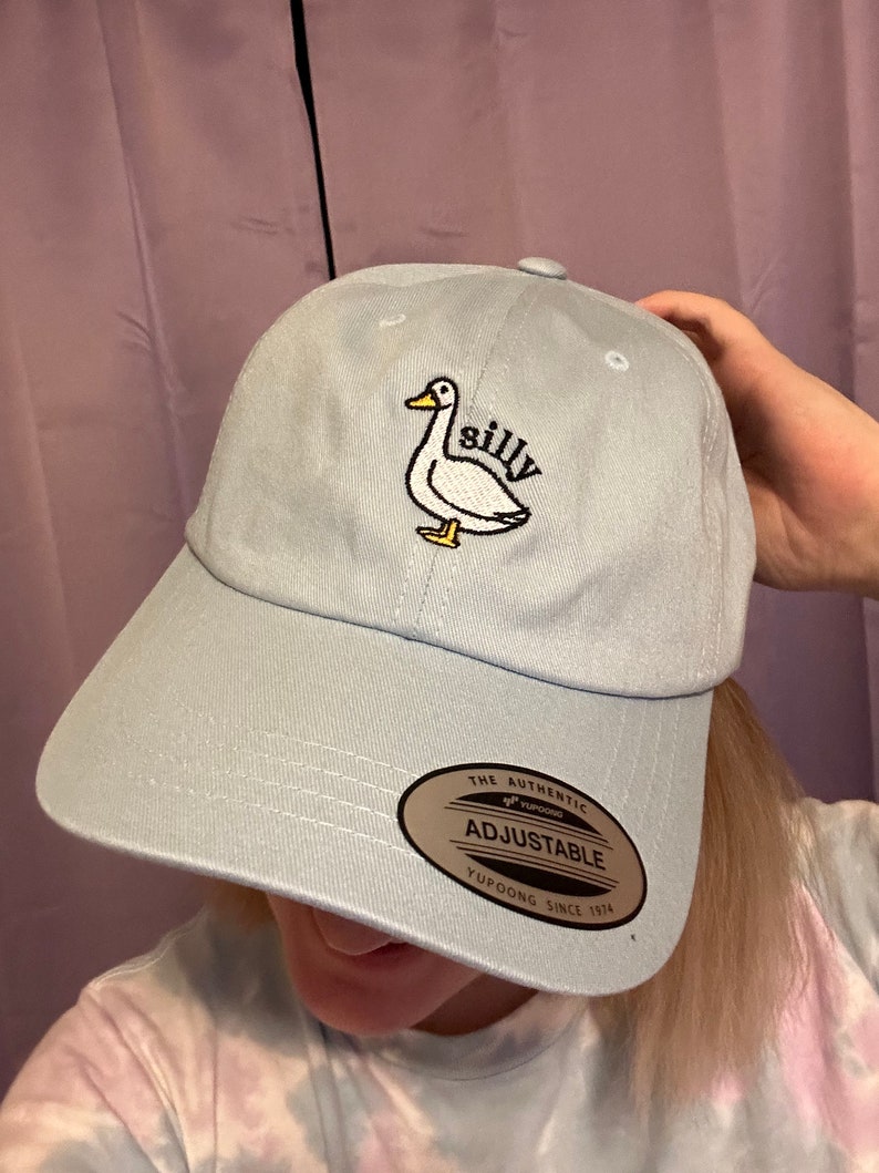 May include: A light blue baseball cap with an embroidered white duck and the word "silly" on it. The cap has an adjustable strap with a brown label that says "The Authentic Yupoong Adjustable Yupoong Since 1974."
