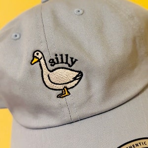 May include: A light blue baseball cap with an embroidered white duck and the word "silly" in black. The cap has an adjustable strap with a black label that says "The Authentic YUPOONG ADJUSTABLE".