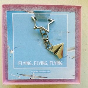 May include: A silver keychain featuring a star charm and a paper aeroplane charm. The keychain is presented in a pink box with a blue background and the text "FLYING, FLYING, FLYING" and "LilyfieldCreations.com".