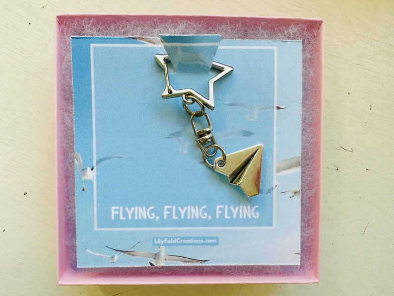 May include: A silver keychain featuring a star charm and a paper aeroplane charm. The keychain is presented in a pink box with a blue background and the text "FLYING, FLYING, FLYING" and "LilyfieldCreations.com".