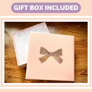 May include: A pink gift box with a gold bow on the lid. The box is sitting on a wooden surface. "Gift Box Included" is written in purple text on a light pink background.