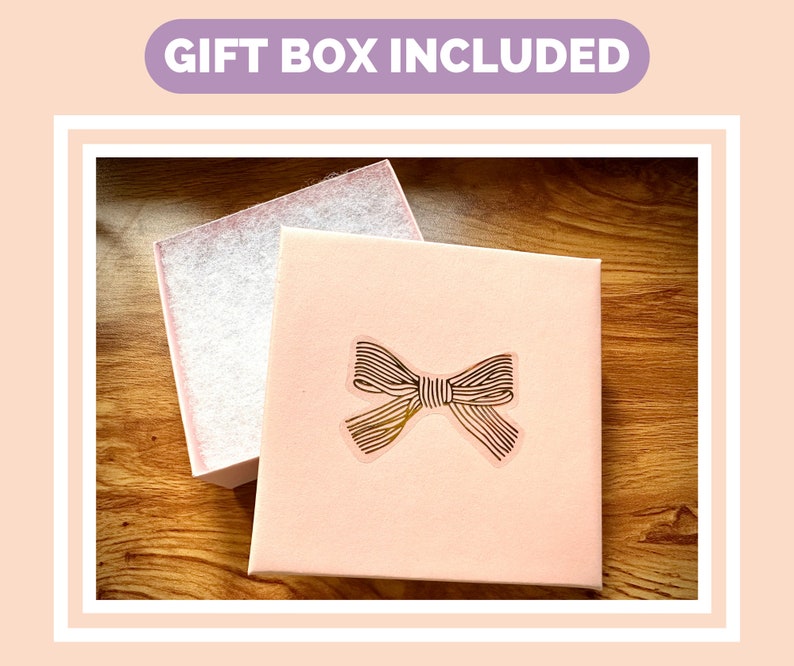 May include: A pink gift box with a gold bow on the lid. The box is sitting on a wooden surface. "Gift Box Included" is written in purple text on a light pink background.