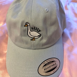 May include: A light blue baseball cap with an embroidered white goose and the word "silly". The cap has an adjustable strap with a black label that reads "The Authentic Yupoong Adjustable Yupoong Since 1974".