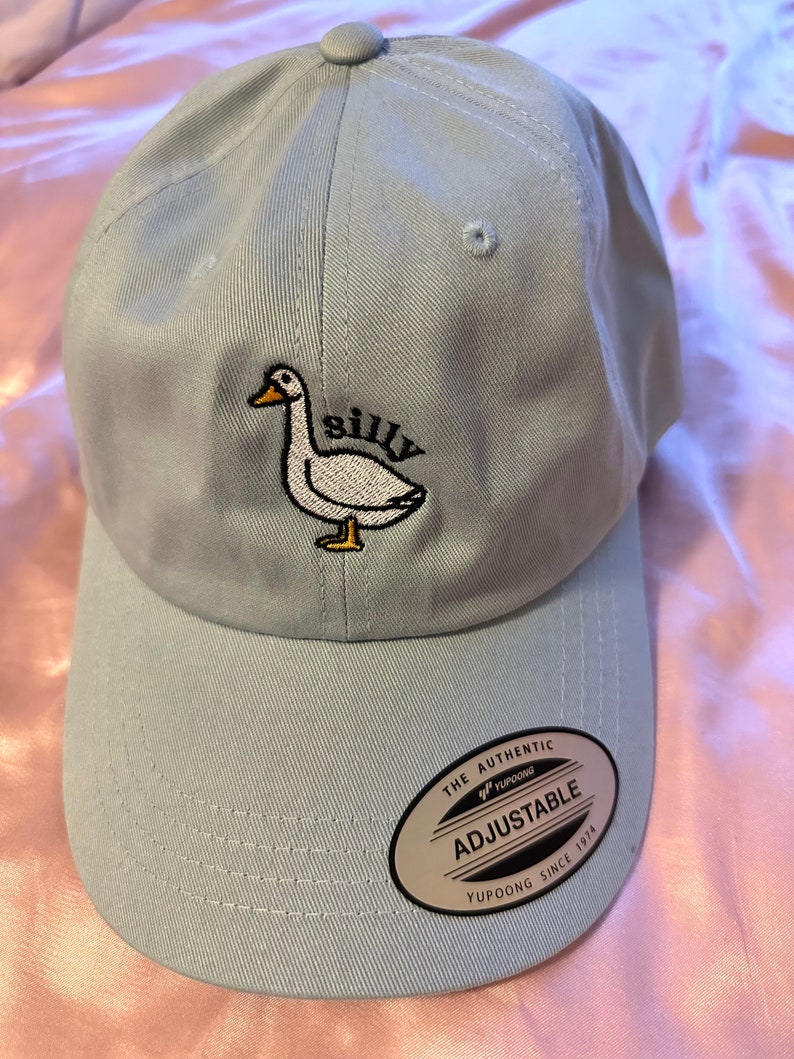 May include: A light blue baseball cap with an embroidered white goose and the word "silly". The cap has an adjustable strap with a black label that reads "The Authentic Yupoong Adjustable Yupoong Since 1974".