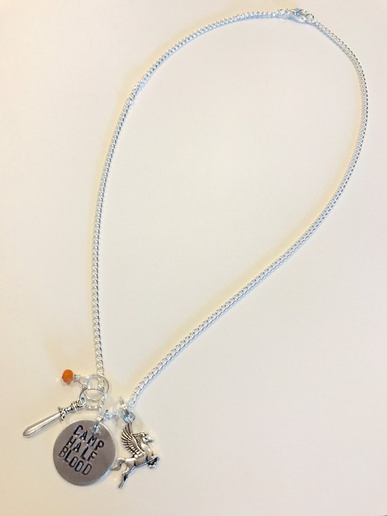 Pode incluir: Um colar de prata com um pingente que diz "Camp Half Blood" e que apresenta um cavalo alado, uma espada e uma conta laranja.