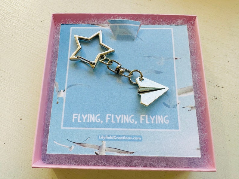 May include: A silver keychain featuring a star and a paper aeroplane charm. The keychain is presented against a blue background with the text "Flying, Flying, Flying" and the website "LilyfieldCreations.com".