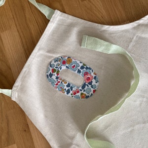 May include: A white canvas apron with a green strap and a floral fabric patch in the shape of the letter "O".