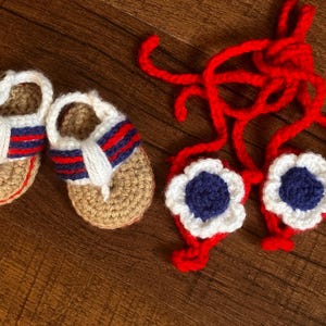 May include: A set of crocheted baby sandals and a headband. The sandals are tan with red, white, and blue stripes. The headband is red with white and blue flowers.