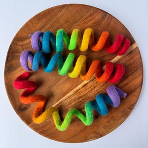 May include: A wooden plate holds six colorful felt spirals in a rainbow of hues. The spirals are arranged in a circular pattern, with each one transitioning from red to orange, yellow, green, blue, and purple. The felt has a soft, textured appearance.
