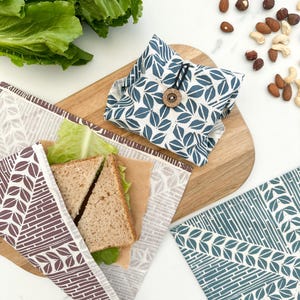 May include: Three reusable food wraps with different leaf patterns. The wraps are made of fabric and are designed to be used as an alternative to plastic wrap. The wraps are shown on a wooden cutting board with a sandwich wrapped in one of the wraps.