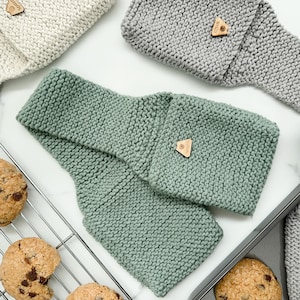 May include: A sage green knitted headband with a small wooden tag. Other knitted headbands in light grey and off-white are visible. Chocolate chip cookies are on a cooling rack and baking sheet.