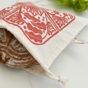 May include: A white cotton bread bag with a red floral design. The bag is tied with a white drawstring and has a small brown tag that says "eco goods".