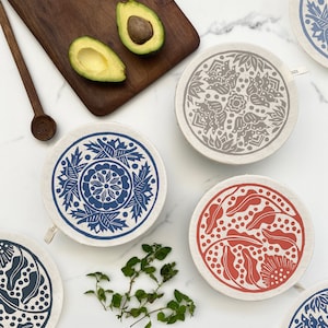 May include: Set of five reusable food wraps with different floral patterns. The wraps are made of fabric and have a circular shape. The patterns are in shades of blue, gray, and red.