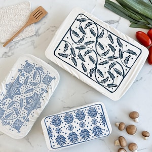 以下が含まれることがあります： 青い花柄の白い長方形のキッチンタオル3枚。1枚は葉のデザイン、もう1枚は花と葉のデザイン、3枚目は花柄です。木製のヘラと野菜も写っています。