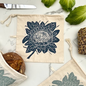 May include: Cream-colored reusable bread bags with a dark teal floral print. The bags have a drawstring closure and a small tag that says "spaza". A loaf of bread is visible in one bag. A knife and lettuce are in the background.