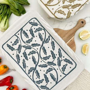 Könnte beinhalten: Ein weißes Geschirrtuch mit einem dunkelblauen Blumenmuster. Das Muster zeigt stilisierte Blätter und Ranken. Das Geschirrtuch ist gefaltet und liegt auf einer weißen Oberfläche.