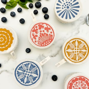 May include: Six reusable food covers with different patterns. The covers are made of fabric and have a white background with colorful designs. The designs include a yellow flower, a blue sunburst, a red floral pattern, a yellow floral pattern, a blue floral pattern, and a pink floral pattern.